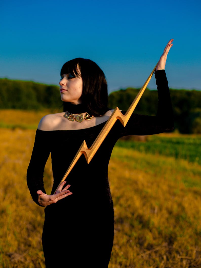 May include: A wooden lightning bolt prop with a gold finish. The prop is held up against a backdrop of a field and blue sky. The lightning bolt is approximately 30 inches long.