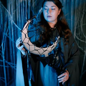 May include: A crescent moon-shaped dreamcatcher with a wooden frame and intricate black webbing. The dreamcatcher features black feathers and a central decorative element. The design is accented with black leather and is held in front of a blue-toned background.