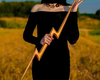 May include: A wooden lightning bolt with a natural wood finish. The bolt is held by a person wearing a black dress. The background features a field of yellow grass and a blue sky.