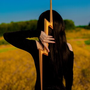 May include: A golden lightning bolt-shaped accessory. The accessory is held against a person's back, with the person wearing a black off-the-shoulder top. The background features a field of yellow flowers and a blue sky.