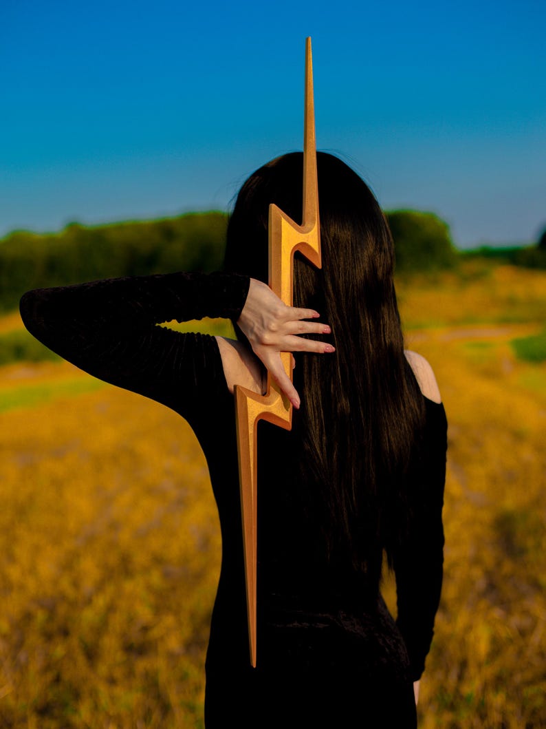 May include: A golden lightning bolt-shaped accessory. The accessory is held against a person's back, with the person wearing a black off-the-shoulder top. The background features a field of yellow flowers and a blue sky.