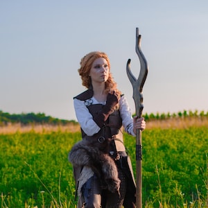 Könnte beinhalten: Eine Person in einer braunen Lederweste, einem weißen Hemd und einem Fellakzent, die einen Holzstab mit geschwungenem Oberteil hält. Die Person hat rotes Haar und weiße Gesichtsbemalung und steht auf einem grünen Grasfeld unter blauem Himmel.