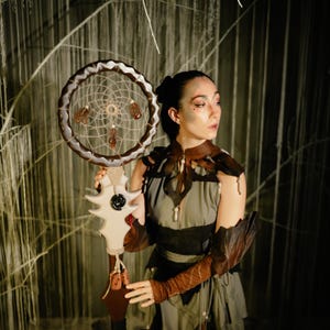 May include: A handcrafted dreamcatcher with a circular woven web and decorative feathers. The dreamcatcher is held by a person wearing a dark green dress and brown leather accents. The background features a textured, neutral-toned backdrop.