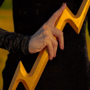 May include: A golden lightning bolt prop with a textured surface. The prop is held against a black background, with a glimpse of a person's arm and clothing. The bolt's design is sharp and angular, with a metallic sheen.