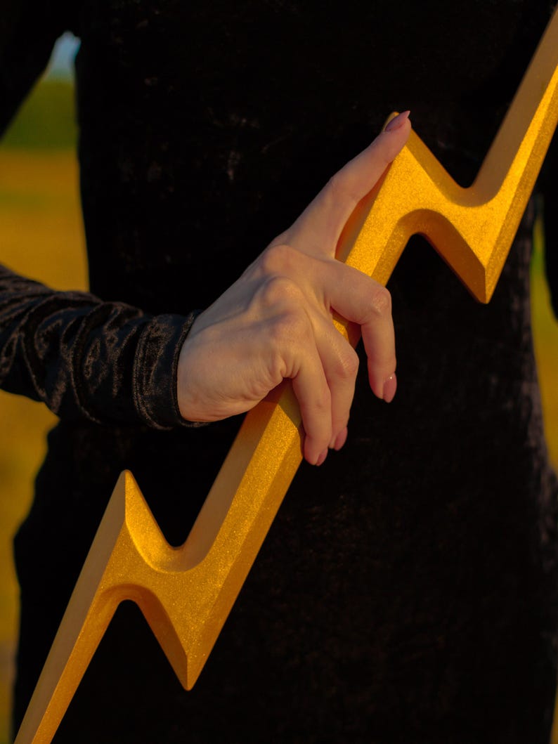 May include: A golden lightning bolt prop with a textured surface. The prop is held against a black background, with a glimpse of a person's arm and clothing. The bolt's design is sharp and angular, with a metallic sheen.