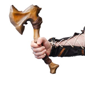 Op de afbeelding: Een houten bijl met een lichtbruine afwerking, met een gebogen blad en een handvat omwikkeld met bruin leer. De bijl wordt in een hand gehouden, wat het vakmanschap van het wapen laat zien.