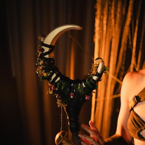 Könnte beinhalten: Ein handgefertigter Stab mit einem halbmondförmigen Oberteil. Der Stab ist mit schwarzem Band, grünen Blättern und roten Pilzakzenten verziert. Der Stab hat einen dunkelbraunen Griff und einen Pilz am unteren Ende.