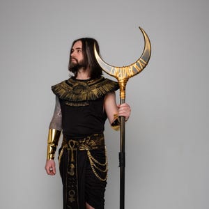 Peut inclure: Un costume sur le thème égyptien avec un personnel doré et noir en forme de croissant de lune. Le costume comprend un haut noir, une jupe et des accents dorés. Le personnel mesure environ 152 cm de haut. La personne porte des bracelets de bras dorés.