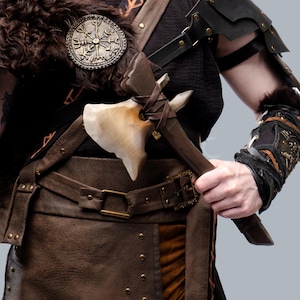 May include: A detailed view of a Viking costume, featuring a brown leather belt, a decorative bronze emblem, and a hand-held axe with a light-colored stone blade. The costume includes fur accents and leather armor.