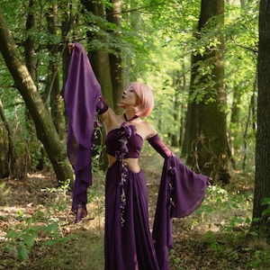 May include: A long, deep purple dress with flowing sleeves and a matching crop top, decorated with floral details. The outfit is worn in a forest setting. The model has pink hair and is posing.