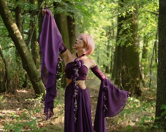 May include: A flowing, deep purple dress with long, draped sleeves and a matching crop top. The outfit is adorned with floral accents and is worn in a forest setting. The model has pink hair.