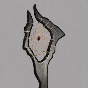 May include: A dark wooden sculpture featuring a dreamcatcher design. The sculpture has a central web with a red bead, held within a frame shaped like horns. The frame is bound with twine, creating a unique, handcrafted aesthetic.
