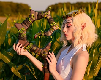 Może przedstawiać: Brązowo-zielona laska fantasy z okrągłym wierzchołkiem, owinięta ciemnobrązowym materiałem i ozdobiona małymi zielonymi listkami. Laska ma długi, czerwonobrązowy uchwyt. Obraz jest ustawiony na zewnątrz z tłem pola kukurydzy.