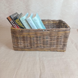 May include: A brown wicker basket with a woven pattern holds a stack of five books. The books are all light blue with white text. The text on the books reads "Атлант" and "Ария Грэй".