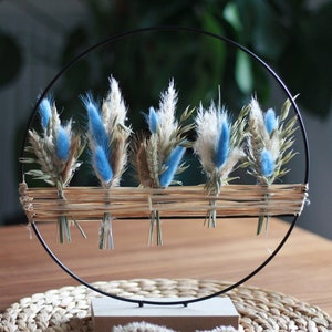 Peut inclure: Cinq petits bouquets de fleurs séchées, dont du pampas bleu et blanc, sont disposés dans un cercle en métal noir. Le cercle est monté sur un socle en bois et repose sur un dessous de plat en paille tressée.