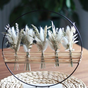 Peut inclure: Un cercle en métal noir avec une base tissée contient cinq petits bouquets de pampas séchés blancs et de blé. Le cercle est posé sur un dessous de plat en paille tissée.