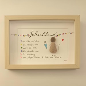 May include: Framed artwork with the word "Schulkind" and a stick figure made of pebbles. The artwork includes colorful text and a small banner. The frame is made of light-coloured wood. The year 2024 is written in the bottom right corner.
