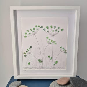 May include: A framed artwork featuring a green sea glass floral design. The artwork is set against a white background and framed in a white frame.