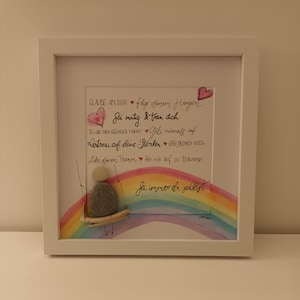 May include: A framed piece of art with a watercolour rainbow and a pebble person on a swing. The art features a handwritten message that reads: "GLAUBE AN DICH folge deinem Herzen Sei mutig & trau dich  TUE WAS DICH GLÜCKLICH MACHT Gibs niemals auf  VERTRAUE AUF DEINE STÄRKEN GEH DEINEN WEG  LEBE DEINEN TRAUM Hör nie auf zu träumen  Sei immer du selbst."