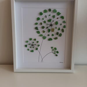 May include: A framed artwork featuring three green glass dandelion designs on a white background. The artwork is signed "Ramona".