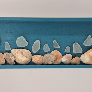 Puede incluir: Una obra de arte enmarcada que presenta un grupo de pequeños pájaros azul claro hechos de vidrio de mar, posados ​​en varias conchas de mar beige y marrones. Los pájaros están dispuestos en una fila sobre un fondo de madera azul.