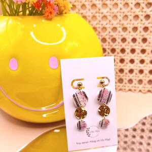 Könnte beinhalten: Ohrringe zum Aufhängen mit rosa, schwarzen und goldenen Akzenten. Die Ohrringe haben geometrische Formen und kreisförmige, goldfarbene Charms. Sie werden auf einer weißen Karte mit Text präsentiert, mit einer gelben Smiley-Vase und Blumen im Hintergrund.