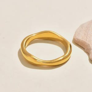 Peut inclure: Une bague dorée au design ondulé. La bague est circulaire et repose sur une surface claire. Une pierre texturée rose clair est en arrière-plan.
