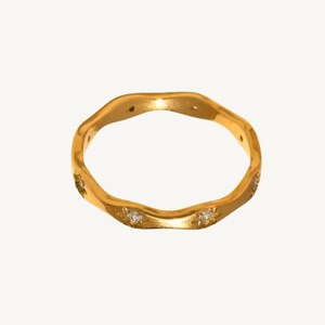 May include: A gold ring with a wavy, undulating band design. The ring features small, clear, round-cut stones embedded at intervals along the outer surface of the metal band, creating a subtle sparkle against the polished gold finish.
