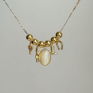 Könnte beinhalten: Eine goldene Kettenkette mit einem Charm-Armband mit einem Hufeisen, einem Cowboystiefel, einem Kaktus und einem Hand-Charm. Das Charm-Armband hat auch einen großen ovalen weißen Stein-Anhänger.