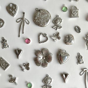 Könnte beinhalten: Eine Sammlung von silberfarbenen Anhängern und Charms in verschiedenen Formen, darunter Herzen, Blumen, Schmetterlinge und Schleifen. Einige Charms weisen kleine farbige Edelsteine auf. Die Charms sind auf einer weißen Oberfläche verstreut.
