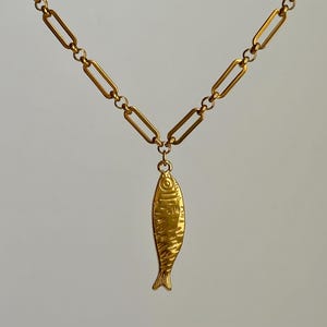 Op de afbeelding: Goudkleurige ketting met een vishanger. De hanger heeft details van schubben en vinnen. De ketting is gemaakt van langwerpige rechthoekige schakels. De ketting wordt getoond tegen een effen achtergrond.