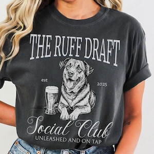 May include: Dark gray t-shirt with a graphic of a dog, a beer glass, and the text "THE RUFF DRAFT SOCIAL CLUB". The design includes the text "est 2025" and "UNLEASHED AND ON TAP".