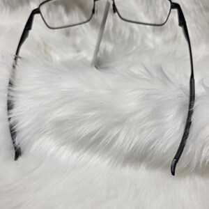 Puede incluir: Gafas rectangulares de marco negro con lentes transparentes. Las gafas est&aacute;n sobre una superficie blanca y esponjosa.