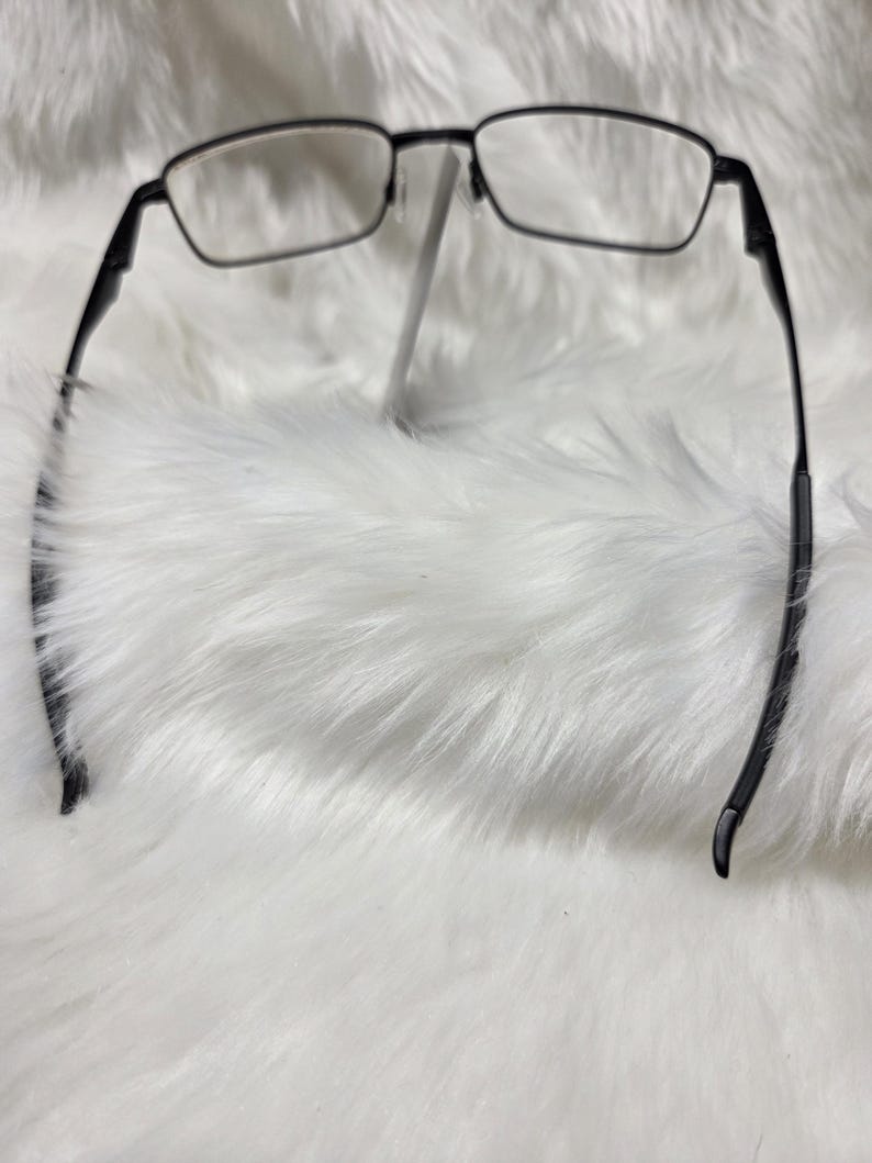 Puede incluir: Gafas rectangulares de marco negro con lentes transparentes. Las gafas est&aacute;n sobre una superficie blanca y esponjosa.