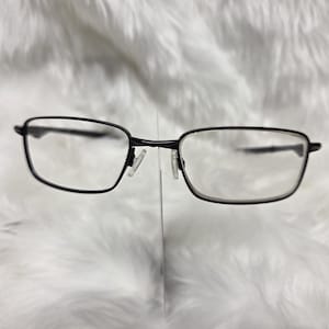 Puede incluir: Gafas rectangulares negras con un fino marco de metal. Las gafas est&aacute;n sobre una superficie blanca y peluda.