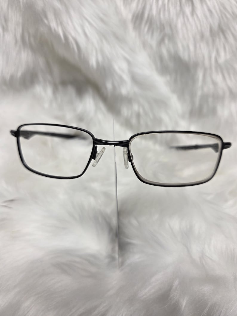 Puede incluir: Gafas rectangulares negras con un fino marco de metal. Las gafas est&aacute;n sobre una superficie blanca y peluda.
