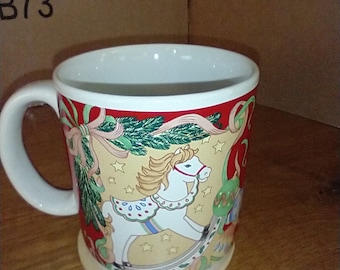 May include: White ceramic mug with a Christmas-themed design featuring a rocking horse, green pine branches, red ribbons, and gold stars. The mug has a white handle.