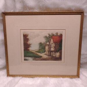 May include: A framed print of a watercolor painting depicting a quaint cottage with a red roof and a stone facade. The cottage is situated on a winding path leading to a small stream. The painting is titled "A Home on the Hill" and is signed by the artist.