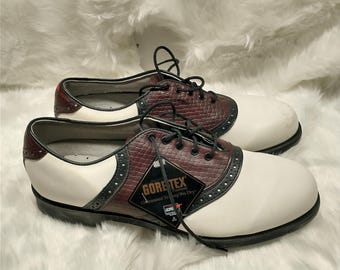 May include: White and brown leather golf shoes with black accents and a Gore-Tex label. The shoes have laces and a saddle design.