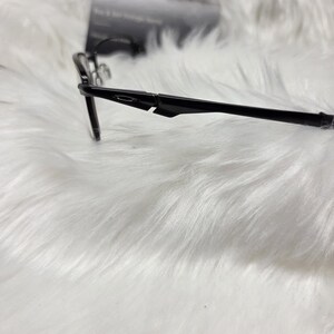 Puede incluir: Gafas negras con un fino marco de metal y patillas de pl&aacute;stico negro. Las patillas tienen una curva sutil y el nombre de la marca es visible en el lateral.