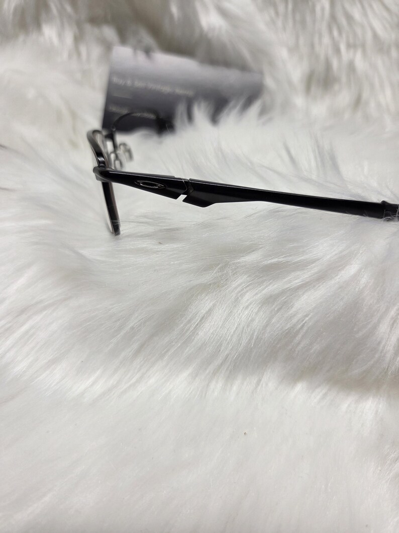 Puede incluir: Gafas negras con un fino marco de metal y patillas de pl&aacute;stico negro. Las patillas tienen una curva sutil y el nombre de la marca es visible en el lateral.