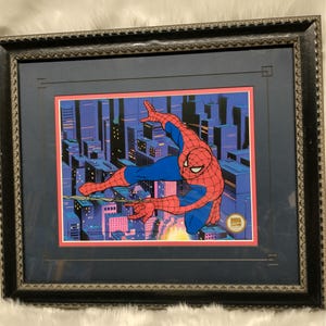 May include: Framed illustration of Spider-Man swinging through a city skyline at night. The image is in a black frame with a blue mat. The image is signed "Marvel Comics" in the bottom right corner.