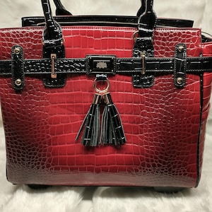 May include: A red handbag with a crocodile skin texture. The bag has black accents, including the handles, straps, and a decorative buckle with a lotus flower emblem. A tassel hangs from the front. The bag is on a white, fluffy surface.