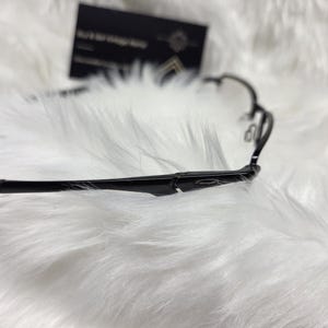 Puede incluir: Gafas negras con un fino marco de metal. Las gafas est&aacute;n sobre una superficie blanca y esponjosa.