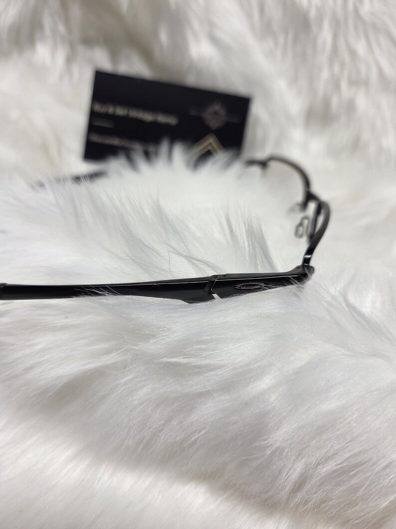 Puede incluir: Gafas negras con un fino marco de metal. Las gafas est&aacute;n sobre una superficie blanca y esponjosa.