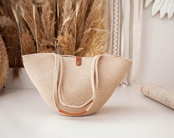 Pode incluir: Uma grande bolsa tote bege, tecida com alças de couro. A bolsa é feita de fibras naturais e tem uma parte superior larga e aberta.