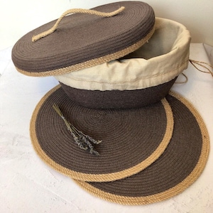 May include: A brown woven basket with a matching lid and two placemats. The basket has a beige interior and a rope handle on the lid. The placemats and the lid have a beige border. A small bundle of dried lavender rests on the placemats.