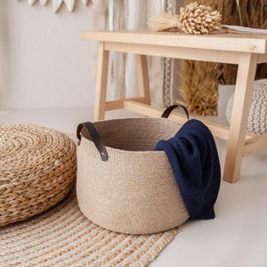 May include: A round, woven jute basket with brown leather handles, holding a navy blue blanket. The basket sits on a natural fiber rug. A woven footstool and wooden bench are in the background, creating a cozy, neutral-toned interior.