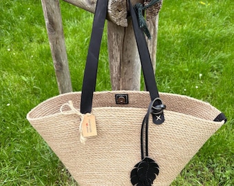 May include: A beige woven tote bag with black leather handles and a black leaf-shaped charm. The bag is hanging on a wooden post in a grassy area. A small tag is attached to the bag.