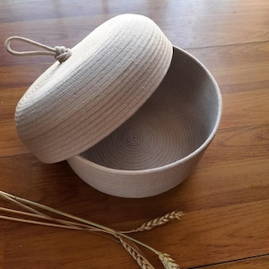 May include: A beige rope bowl with a matching lid, partially open. The bowl and lid are made of tightly wound rope, creating a textured surface. A rope handle is attached to the lid. The bowl sits on a wooden surface, with dried wheat stalks in the foreground.